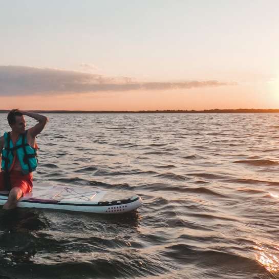 Man sitting on SUP paddleboard with lifevest on and sunset